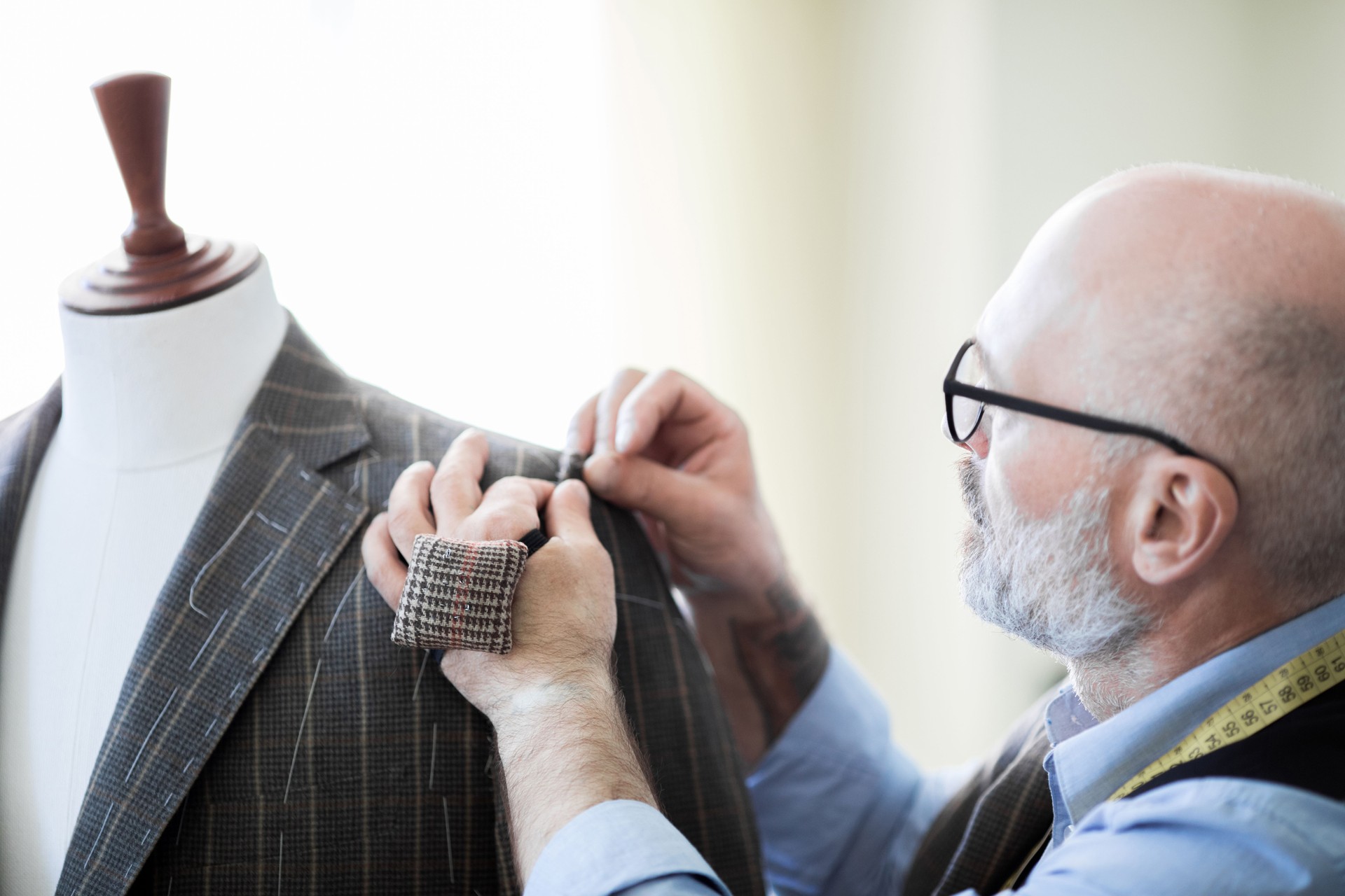 Close-up of concentrated handsome bald tailor with beard wearing glasses holding fabric together with sewing pins while creating design for suit in workshop